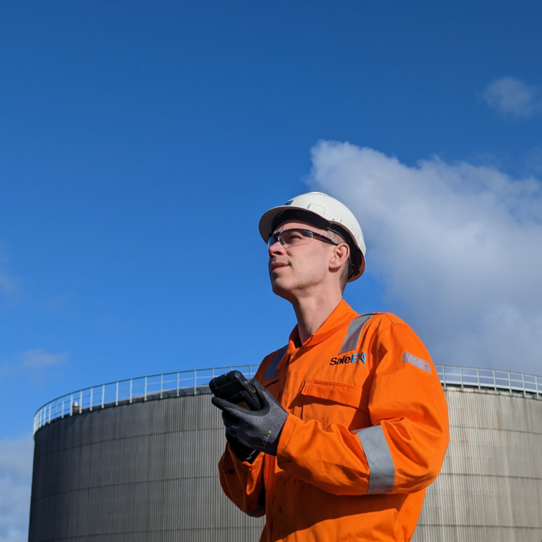 SafeEx Ex technician in orange protective clothing holding a mobile device while working outdoors at an industrial facility.